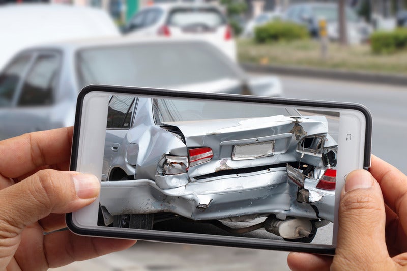 A person holds a smartphone displaying a photo of a damaged car, highlighting the extent of the vehicle's injuries.