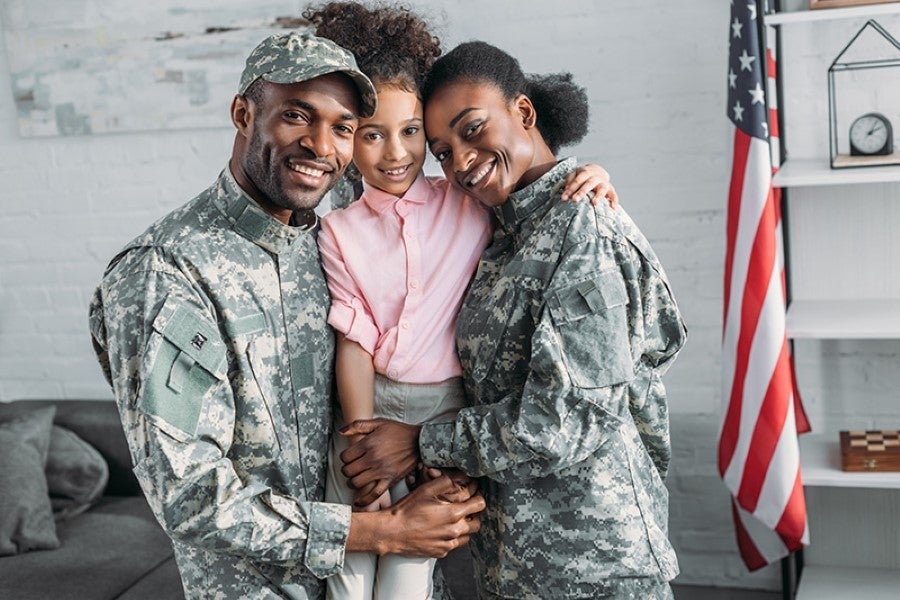 A military family stands together smiling in front of a large American flag, showcasing pride and unity.