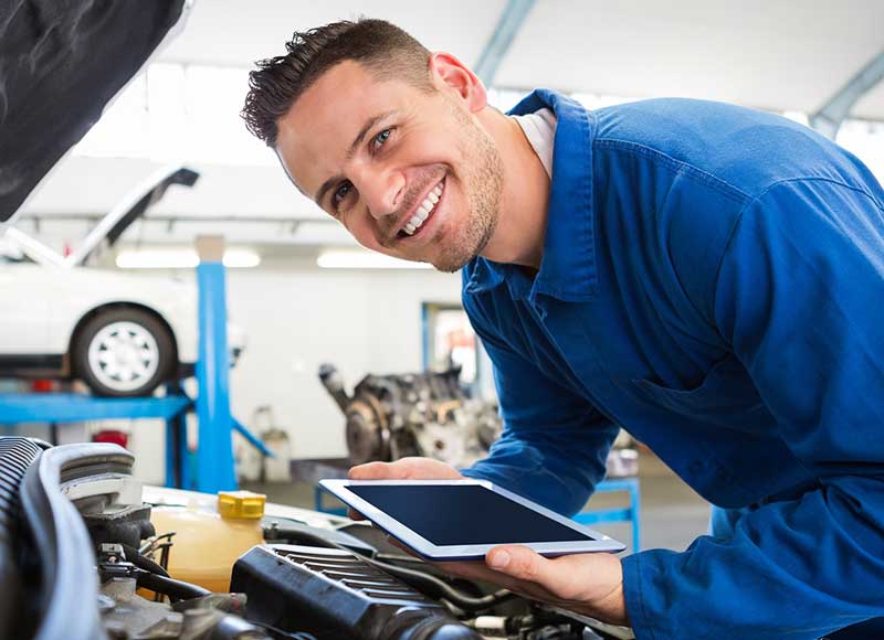 A man examines his car while holding a tablet, focused on the device in his hands.