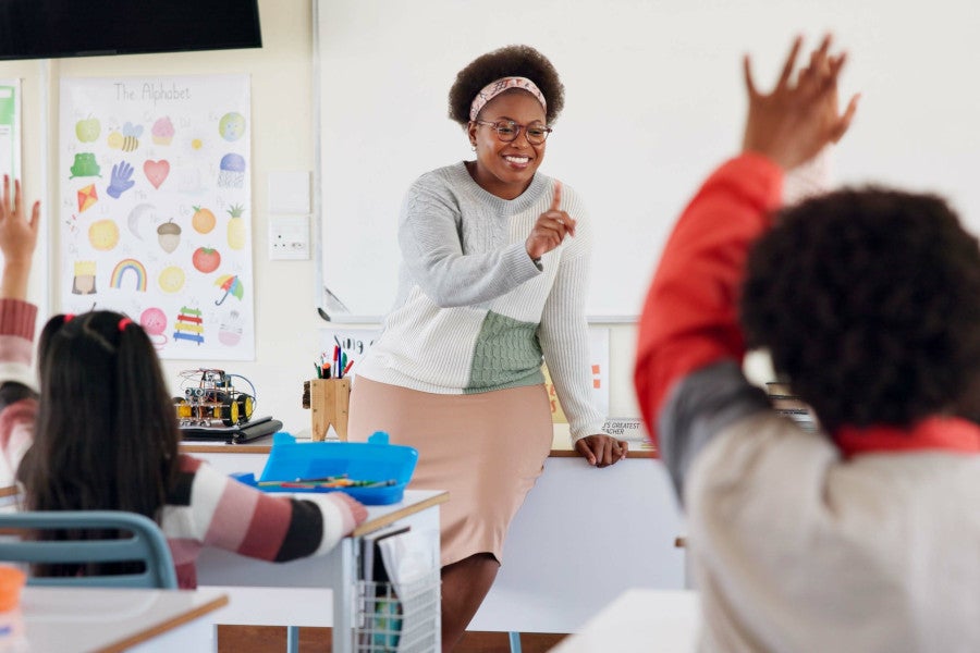 A teacher with raised hands stands in front of a classroom, engaging students.